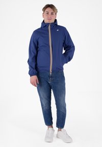 Giacca leggera blu navy con zip frontale, dettagli a strisce bianche e rosse e polsini elasticizzati, abbinata a jeans blu e sneakers bianche.