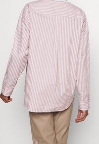 Langærmet skjorte i blødt pink med hvide lodrette striber, har en scallop-kantet krave, sideslidser og knappede manchetter.