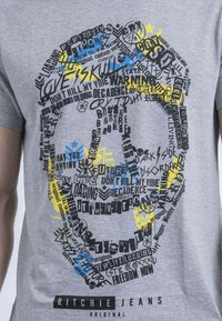 T-shirt en coton gris arborant un grand motif de crâne composé de texte en noir, bleu et jaune. La marque "Ritchie Jeans" est imprimée en dessous.