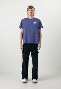 Camiseta de algodón azul con un pequeño gráfico en el lado izquierdo del pecho, combinada con pantalones negros y zapatillas blancas. Diseño simple y relajado.