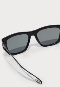 Óculos de sol pretos com lentes cinzentas. Armação de plástico brilhante, design angular, hastes pretas finas e detalhes transparentes nas extremidades das hastes.
