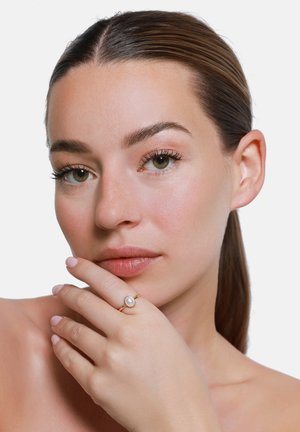 Frau mit heller Haut und braunen Haaren hält eine Hand mit einem goldenen Ring mit einer weißen Perle nahe ihrer Lippen und schaut in die Kamera.