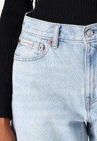 Jeans de mezclilla azul claro con herrajes de cobre, que cuentan con una etiqueta del logo de Calvin Klein en rojo y blanco en el bolsillo frontal y una tela texturizada.