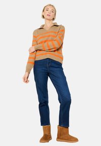 Svetlo rjav pulover z oranžnimi črtami, ovratnikom in rebrastimi manšetami, ki je vparen z temno modrimi ravnimi jeans hlačami in rjavimi gležnjarji.
