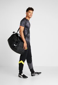 Borsa da viaggio nera con logo, indossata a spalla. Modello in maglietta sportiva grigia texture con dettagli arancioni, pantaloni da corsa neri e gialli e scarpe da ginnastica nere.