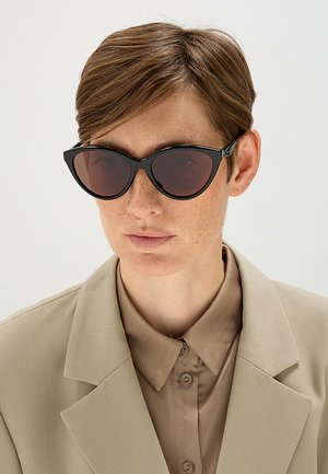 Persona con capelli corti castano chiaro che indossa occhiali da sole scuri a forma di occhio di gatto, una camicia taupe e una giacca beige su uno sfondo semplice.