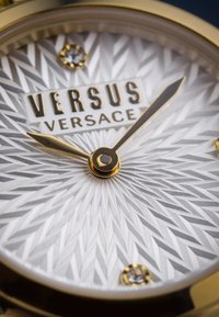 Kultakylkinen kellotaulu, jossa on teksturoitu hopeinen näyttö, jossa on geometrinen kuvio, brändilogo ja kaksi kristallikoristetta. Kultaiset viisarit ja tuntimerkit.