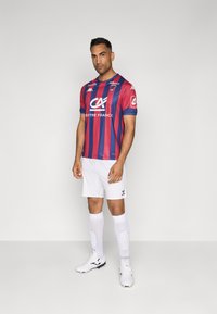 Raudona ir mėlyna dryžuota futbolo marškinėliai su baltais šortais ir kojinėmis. Marškinėliai turi apvalų apykaklę ir logotipus priekyje bei ant rankovių. Avalynė balta.