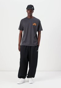 Herretøj bestående af en mørkegrå Nike t-shirt med orange logo, sorte løse bukser og hvide sneakers, suppleret med en sort kasket.
