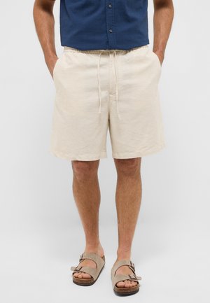Beige Shorts mit elastischem Bund und Kordelzug, gefertigt aus atmungsaktivem Stoff. Getragen mit grauen Wildledersandalen mit verstellbaren Riemen.