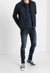 Hombre vestido con chaqueta oscura navy, camiseta polo navy, jeans ajustados desgastados y zapatillas altas navy, de pie con las manos detrás de la espalda.