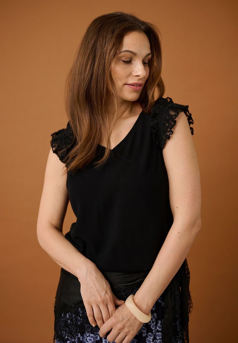 Femme aux longs cheveux bruns portant un haut noir sans manches avec des manches en dentelle et un bracelet beige, regardant vers le bas sur un fond marron.