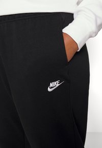 Mustat collegehousut, joissa on sivutaskut ja valkoinen broderattu Nike-logo vasemmassa reidessä. Valmistettu pehmeästä, tekstuurista kankaasta.