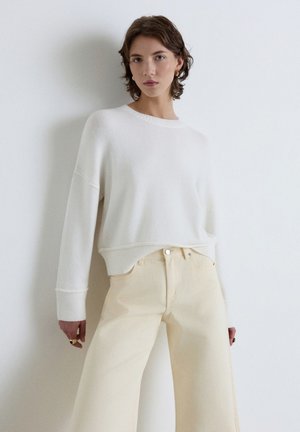 Young woman with short brown hair wearing a loose white sweater and high-waisted cream pants against a plain white background.