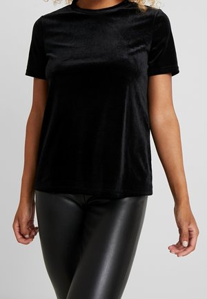 Nainen, joka käyttää lyhythihaista mustaa samettitoppia ja mustia nahkamaista istuvia housuja vaaleanharmaalla yksinkertaisella taustalla.