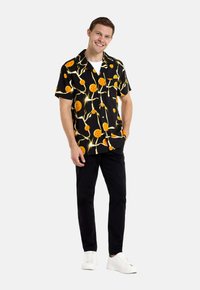 Camisa negra de manga corta con un patrón de frutas naranjas y ramas; combinada con pantalones negros y zapatillas blancas, de pie contra un fondo sencillo.