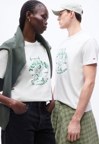 Δύο άτομα κοιτάζονται, φορώντας λευκά t-shirts με το γραφικό "Tommy's Los Angeles"· το ένα έχει ένα πράσινο πουλόβερ περασμένο πάνω από τους ώμους.