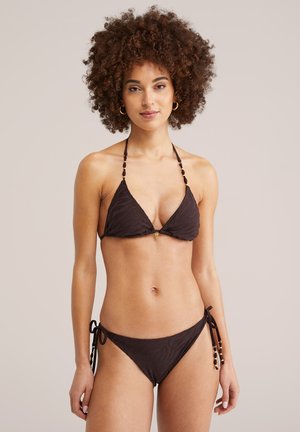 Moteris su garbanotais plaukais, vilkinti tamsiai rudos spalvos raštuotą bikini su aptemptu viršumi su kaklo užsegimu ir šoninėmis rišamomis kelnaitėmis, stovinti priešais paprastą foną.