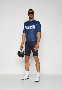 Tmavě modrý cyklistický dres s bílým logem "LE COL", doplněný černými šortkami. Model drží černou přilbu a má na nohou bílé cyklistické boty.