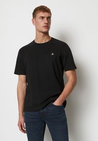 Camiseta de algodón negra con cuello redondo, mangas cortas y un pequeño logo blanco en el pecho. Lleva un par de jeans de mezclilla oscuros.