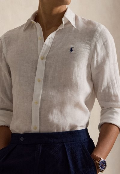 Άνδρας που φοράει λευκό λινό πουκάμισο με κουμπιά και τυλιγμένα μανίκια, σκούρο μπλε παντελόνι και ρολόι χειρός με μπλε καντράν και καφέ δερμάτινο λουράκι.