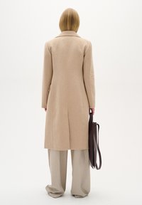 Bēšs vilnas mētelis ar pielāgotu dizainu, garu taisnu griezumu un aizmugures venti. Apvienots ar gaiši pelēkiem platām biksēm un brūnu somu.