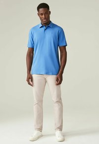 Blaues Polohemd mit Kragen, kurzen Ärmeln und einem dezenten Logo. Kombiniert mit hellbeigen Hosen und weißen Sneakers vor einem neutralen Hintergrund.