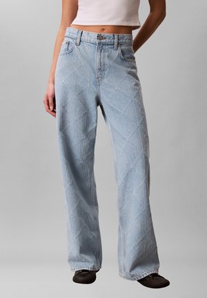 Femme portant un jean large matelassé bleu clair et des chaussures noires, debout les mains le long du corps sur un fond uni.