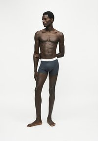 Homme mince debout pieds nus, portant un boxer sombre avec une ceinture blanche, regardant vers la gauche sur un fond blanc uni.
