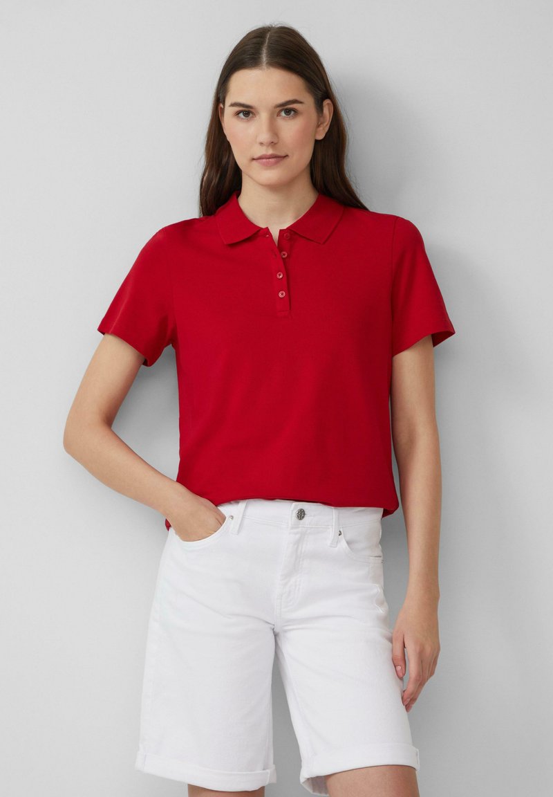 Rode polo met korte mouwen en een kraag, voorzien van een drieknopssluiting, in combinatie met witte denim shorts met omslag.