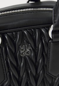 Black leather handbag featuring a braided texture, silver zip closure, and embossed "HUGO" logo. Smooth and glossy surface.
