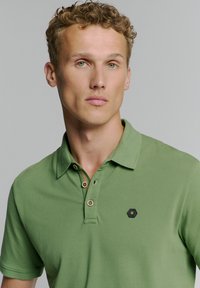 Groene polo gemaakt van katoen, met een kraag, drie-knopenlijst en een hexagonaal logo op de borst. Gestructureerde stof.