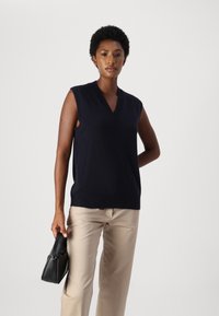 Maglione senza maniche di colore navy con scollatura a V e bordo a costine, abbinato a pantaloni beige. Modella che tiene una piccola borsa nera.