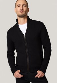 Czarny, dziany sweter zapinany na zamek, z wysokim kołnierzem, solidnym zamkiem błyskawicznym oraz ściągaczami na mankietach i dolnym końcu, noszony na białej koszulce.