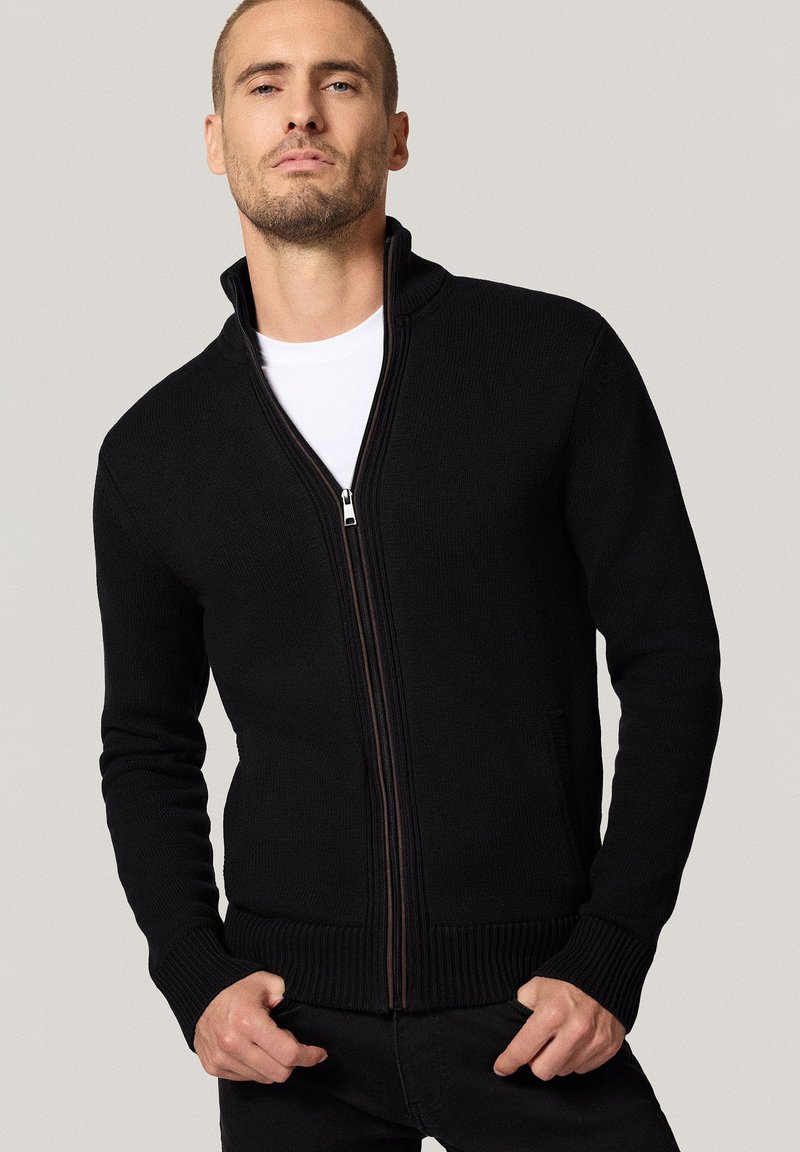 Czarny, dziany sweter zapinany na zamek, z wysokim kołnierzem, solidnym zamkiem błyskawicznym oraz ściągaczami na mankietach i dolnym końcu, noszony na białej koszulce.