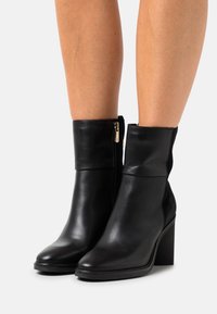 Bottines en cuir noir avec un talon haut cylindrique, texture lisse, fermeture éclair sur le côté et un design de bout effilé.