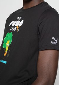 Camiseta de algodón negra con un gráfico colorido de un árbol con ojos de dibujos animados y el texto "EL CLUB PUMA", con un logo de Puma en la manga.