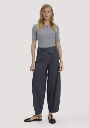 Vrouw die een gestreept topje met korte mouwen draagt, een marineblauwe broek met hoge taille en wijde pijpen met een strikceintuur, en zwarte gewebde platte sandalen, staand.