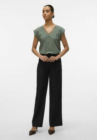 Top verde oliva a maniche corte con dettagli in pizzo, abbinato a pantaloni larghi neri a vita alta e scarpe con tacco scure con accenti.