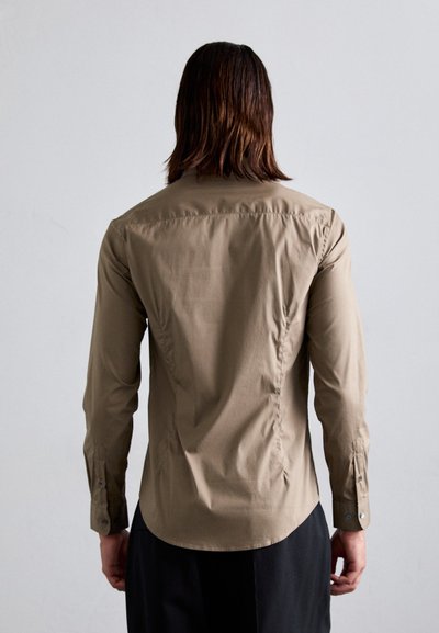 Personne avec des cheveux mi-longs portant un haut ajusté à manches longues beige et un pantalon noir, faisant face à un fond gris clair uni.