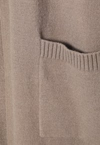 Tissu de pull en tricot beige avec une texture douce, comportant une poche visible et des détails côtelés le long du bord.