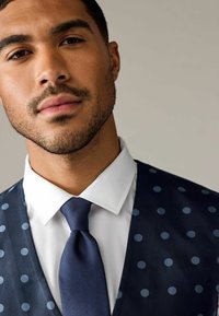 Navy-blaues Polka-Dot-Weste über einem weißen Hemd, kombiniert mit einer strukturierten navyblauen Krawatte. Nahaufnahme, die sich auf Design- und Farbdetails konzentriert.