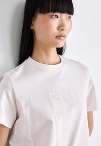 Lys rosa bomull t-skjorte med kort ermer og rund hals, med preget 'Calvin Klein'-logo i gulltråd på forsiden.