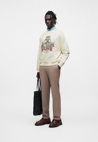 Homme debout portant un sweat crème avec un emblème et un texte, une chemise bleue à col, un pantalon beige, des chaussures marron, tenant un sac fourre-tout noir dans la main droite.