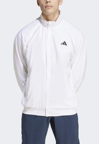 Witte lichte ritsjack met een staande kraag, voorzien van een klein zwart Adidas-logo op de linkerborst en geribbelde manchetten en zoom.