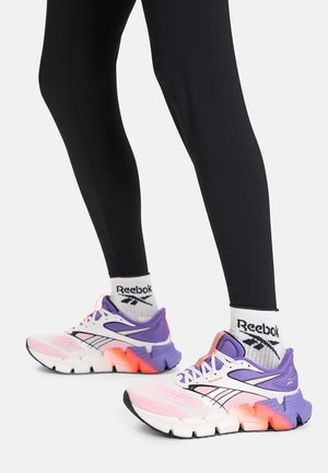 Pies con calcetines blancos de Reebok y zapatillas de correr en tonos rosa, púrpura y blanco con detalles en naranja, combinados con leggings negros.