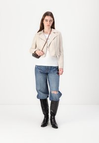 Krémová kožená prešívaná motorkárska bunda, biele tričko, poškodené modré džínsy a čierne vysoké čižmy po kolená. Malá hnedá crossbody kabelka.