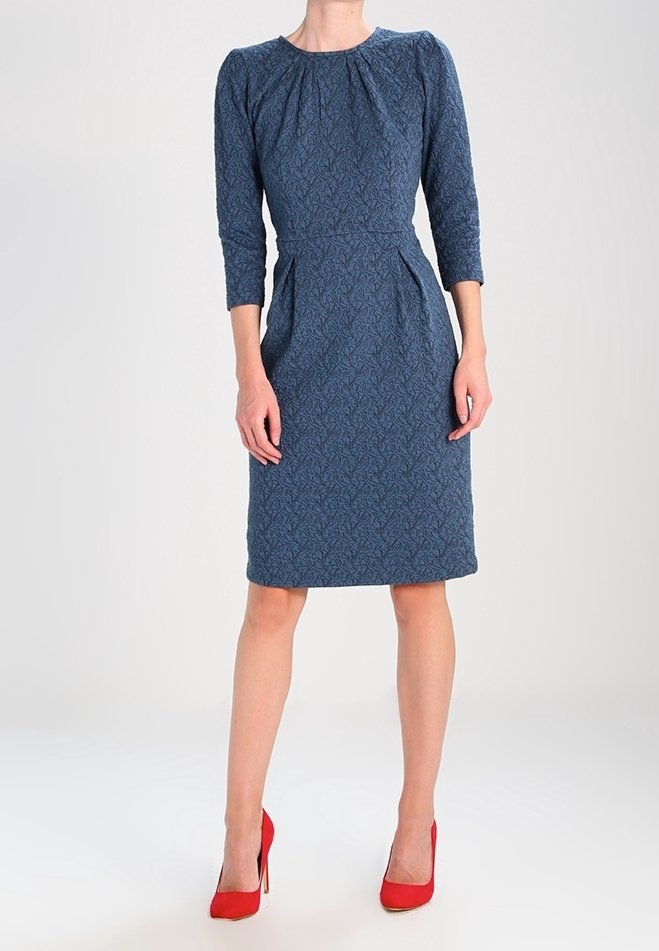 Robe bleue à motifs avec manches trois-quarts, encolure ronde, plis à l'avant et taille cintrée. Associée à des escarpins rouges.