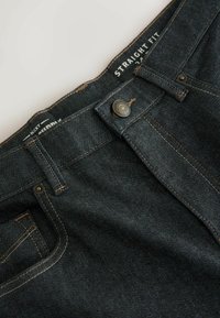 Sötétkék egyenes fazonú farmernadrág, sima textúrával, kontrasztos aranyszínű varrással és fémből készült gombos zárral.