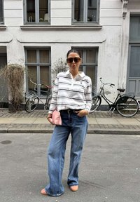 Vrouw met zonnebril, gestreept shirt met lange mouwen, brede jeans, houdt een roze clutch vast, staand op straat met fietsen en een gebouw op de achtergrond.